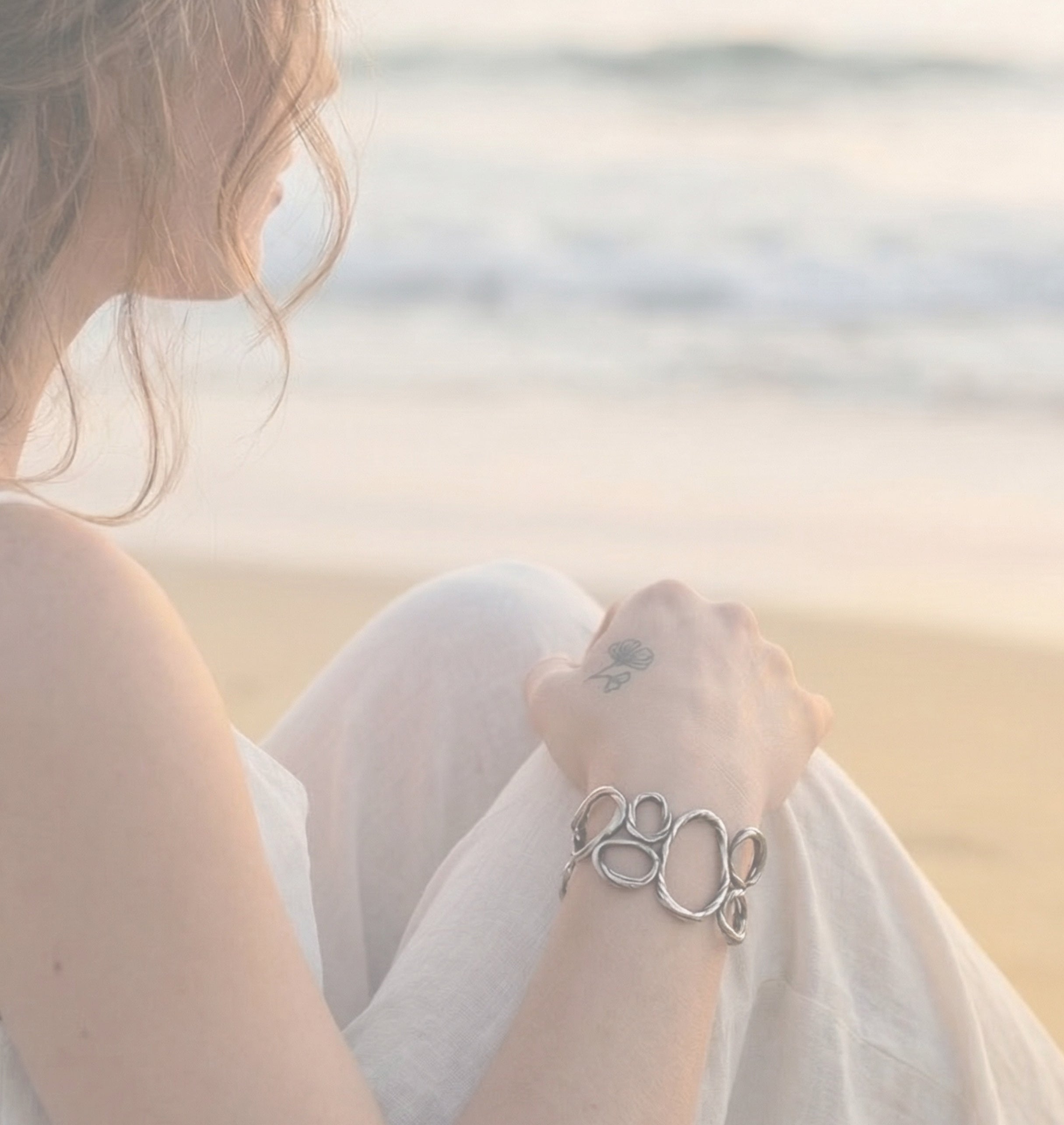 Woman sitting on the beach looking toward the ocean wearing the Pebble Bracelet in sterling silver by jewelry artist Michelle Hoting, inspired by smooth beach stones.