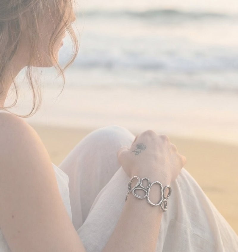 Woman sitting on the beach looking toward the ocean wearing the Pebble Bracelet in sterling silver by jewelry artist Michelle Hoting, inspired by smooth beach stones.