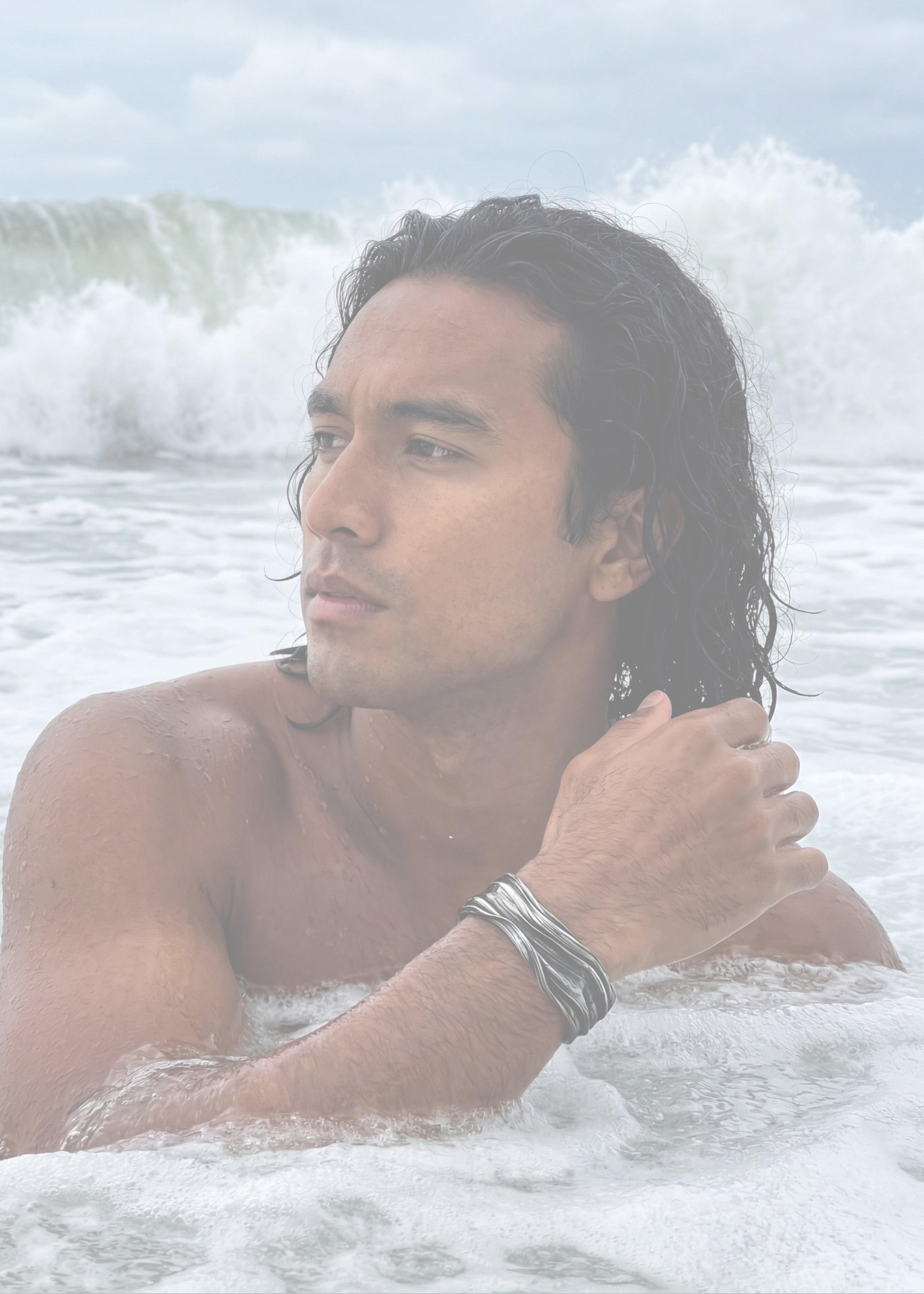 Man standing in the ocean with waves crashing around him wearing the meander cuff bracelet